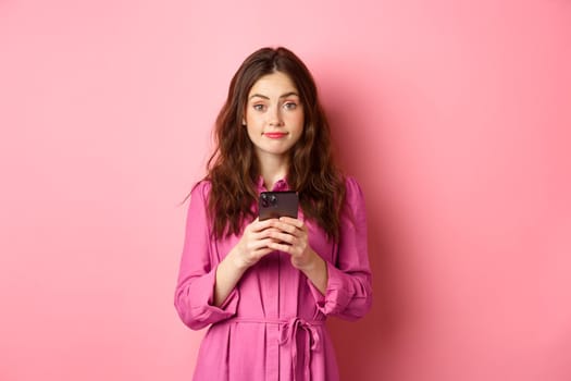 Cute young woman using mobile phone app, holding smartphone and smiling at camera, standing in dress against pink background. Copy space