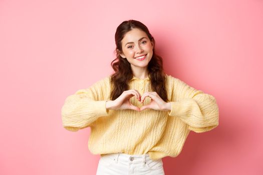 Beautiful romantic girl say I love you, showing heart sign and smiling at camera, standing cute against pink background.