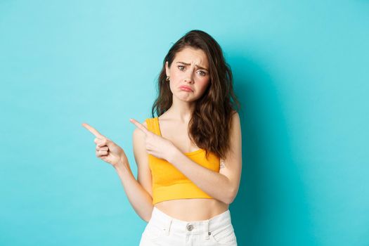 Upset and gloomy brunette girl complaining, pointing fingers left at sad unfair thing, showing bad news, standing against blue background.