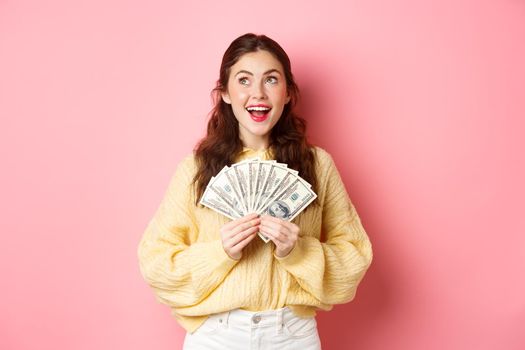 Beautiful happy woman looking dreamy aside, holding dollar bills, thinking of shopping or wasting money, standing against pink background.