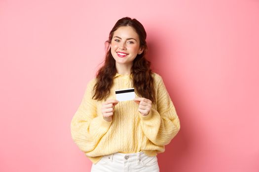 Beautiful female model thinking of shopping, holding plastic credit card and smiling, standing against pink spring background. Copy space