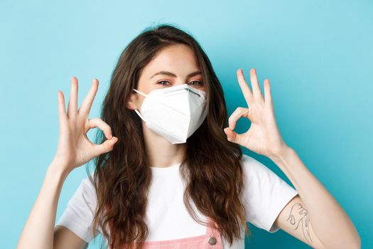 Covid-19, social distancing and lockdown concept. Young positive woman in respirator showing okay signs and nod in approval, praising using preventive measures during pandemic, blue background.