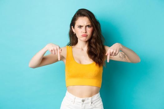 Confused frowning woman looking displeased, need explanation, pointing fingers down at something strange or unpleasant, standing over blue background.