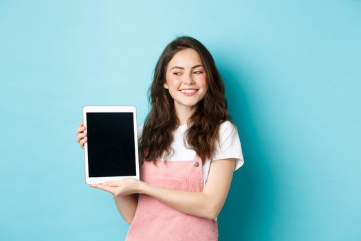 Image of happy young girl showing digital tablet screen and smiling proud, showing your logo on display, standing over blue background.