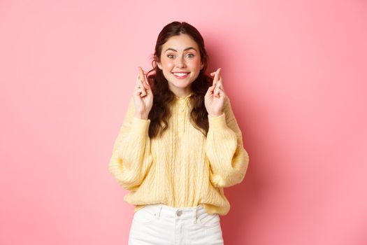 Image of cute cheerful woman cross her fingers for good luck and looking hopeful at camera, making wish, standing over pink background.