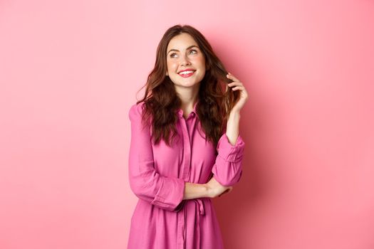 Beautiful coquettish woman in trendy dress and make up, looking thoughtful aside, gazing up with dreamy smile, playing with hair, standing against pink background.