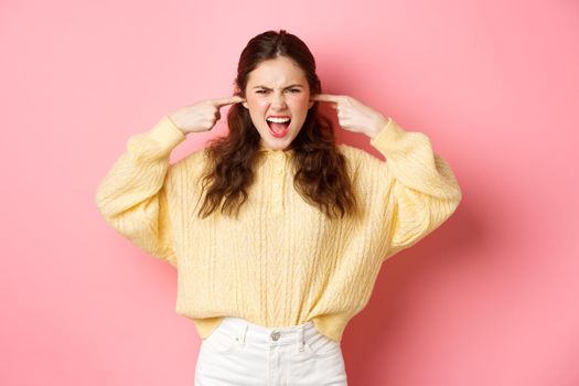 Angry woman blocking her ears with fingers and yelling to turn off music, annoyed by loud noise or disturbing sound, standing against pink background.