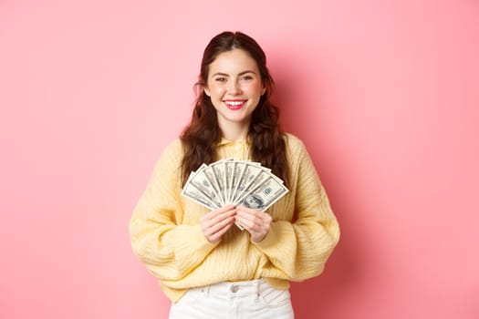 Young happy millennial girl showing dollar bills, holding money with pleased face, winning cash prize, going shopping, standing against pink background.