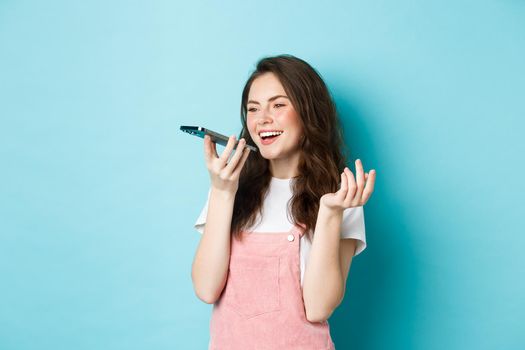 Portrait of smiling woman holding phone near lips and talking, using app translator on smartphone or recording voice message, speaking with speakerphone, standing over blue background.