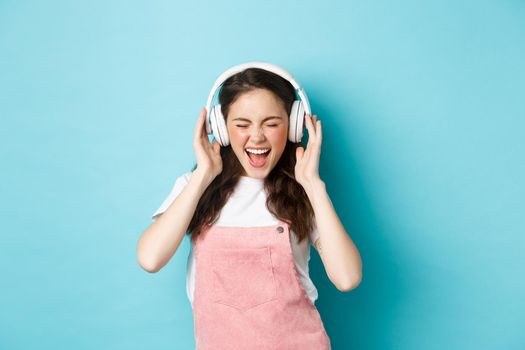 Carefree pretty girl scream from joy, listening music in headphones with happy face, lip-sync lyrics, standing over blue background.