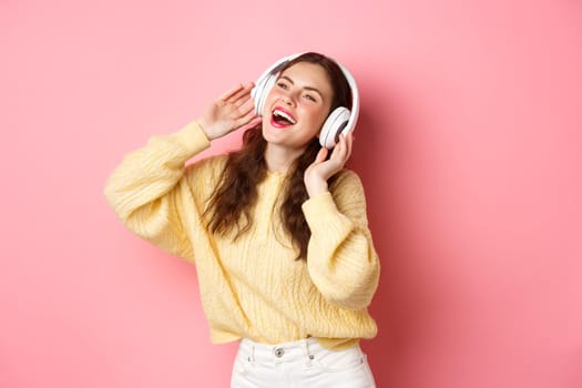 Beautiful modern girl singing favorite song, listening to music in wireless headphones, smiling and dancing, standing over pink background.