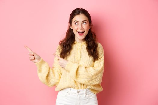 Happy good-looking girl demonstrate information, pointing fingers left and staring at promotional text with pleased smile, standing against pink background.