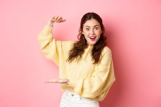 Young happy woman staring at camera, showing big object, holding your logo or product with her hands aside on copy space, standing against pink background.