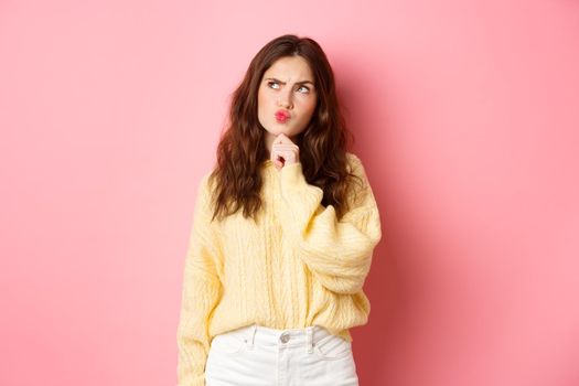 Puzzled cute girl thinking, touching chin and frowning thoughtful, looking aside at logo banner with pensive face, standing over pink background.