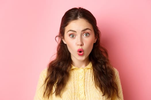 Close up portrait of surprised brunette girl saying wow, folding lips and stare amazed at camera, checking out awesome news, standing against pink background.