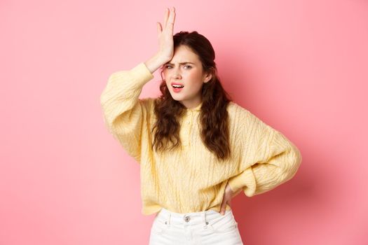 Annoyed and frustrated young woman face palm, holding hand on forehead and staring disappointed at camera, standing against pink background.