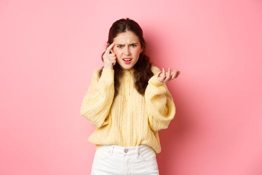 Confused and annoyed girl pointing at head with index finger, scolding someone for stupid or crazy behaviour, being disappointed, standing against pink background.