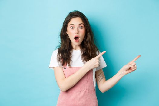 Excited and surprised young woman open mouth, say wow and pointing fingers right at logo, staring impressed at camera while talking about new product or news, blue background.