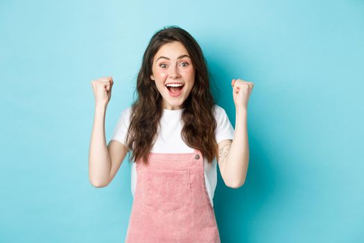 She is winner. Cheerful young woman rejoicing winning prize, achieve goal or success, shouting yes with joyful smile and raising hands up to celebrate triumph, blue background.