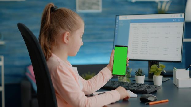 Child vertically holding smartphone with green screen while sitting at desk and doing homework. Little girl looking at isolated background and mockup template for chroma key on display