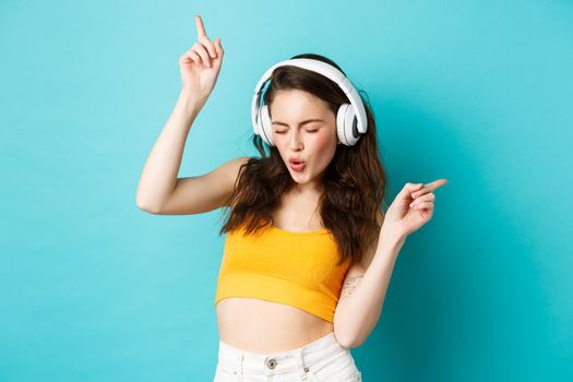 Stylish modern woman in headphones, listening music and dancing, enjoying favorite songs in earphones, standing against blue background.