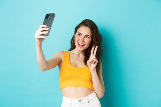 Technology and lifestyle concept. Stylish young woman taking selfie in her summer clothes, showing v-sign at smartphone camera, standing over blue background.