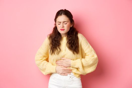 Young woman with pain in stomach, holding hands on belly, feeling terrible ache, menstrual cramps, standing against pink background.