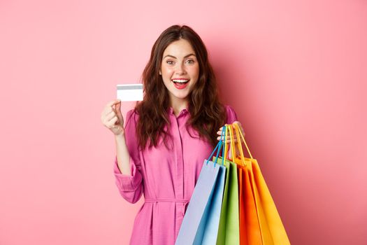 Portrait of excited happy shopaholic, woman holding shopping bags and showing plastic credit card, smiling amazed, standing against pink background.