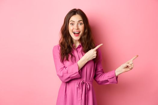 Beauty and fashion. Amazed stylish modern woman pointing fingers right, showing promo on copyspace aside, standing against pink background.