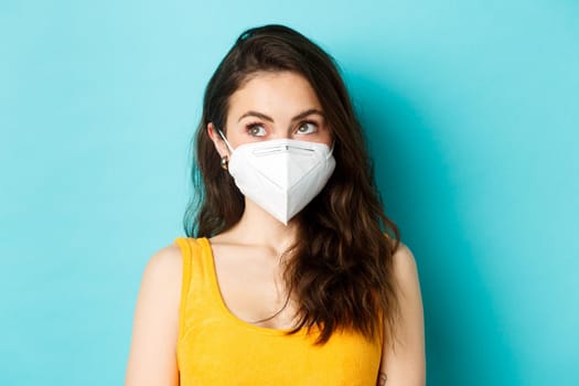 Covid-19, coronavirus and social distancing. Close up portrait of young modern woman in respirator, looking at upper left corner logo, standing against blue background.