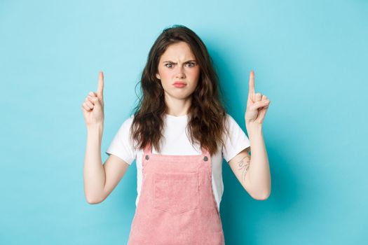Confused and shocked young woman frowning, staring at camera displeased, demand answers, pointing fingers up on top banner, standing over blue background.