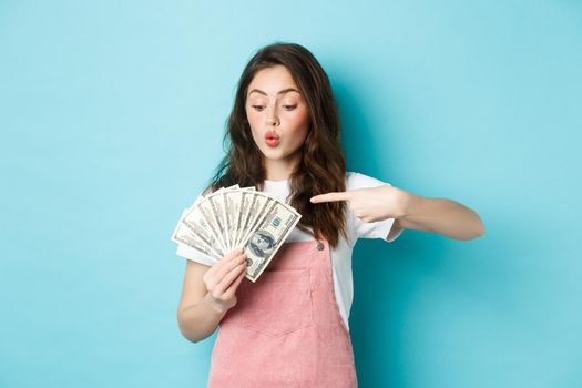 Excited cute girl holding money and pointing at dollar bills, going on shopping, standing over blue background.