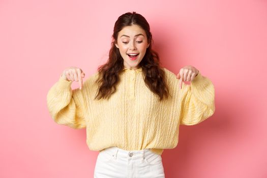 Portrait of happy young brunette woman checking out promo offer, pointing and looking down with amazed face, standing over pink background.