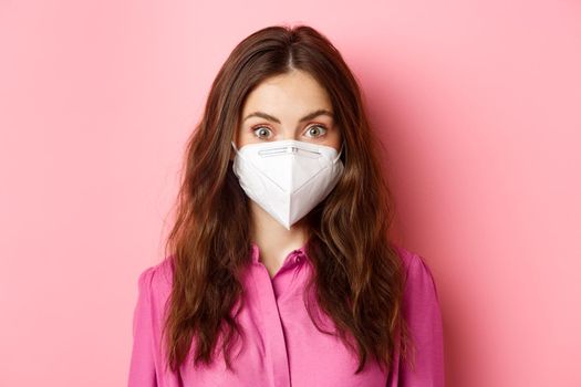 Covid-19, quarantine and social distancing concept. Close up of young stylish woman with nice hairdo, wearing medical respirator from coronavirus, pink background.