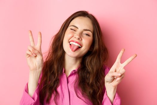 Close up of funny and happy young woman having fun, winking and showing tongue, making v-sign peace gesture, standing over pink background.
