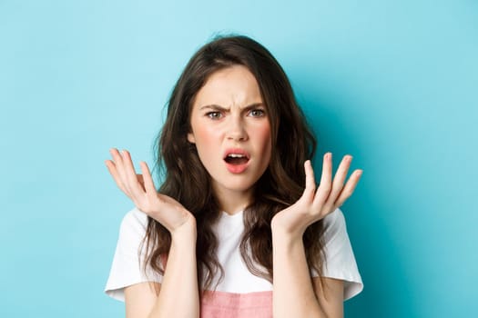 Close up portrait of confused and annoyed brunette girl frowning, grimacing puzzled and shrugging, cant understand, standing over blue background.