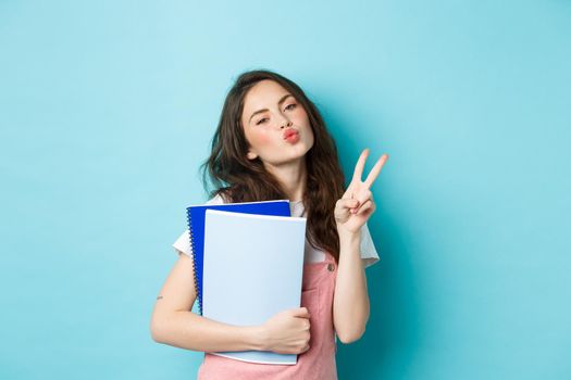 Portrait of glamour girl showing kissing face and v-sign, carry notebooks homework material, standing over blue background.