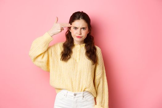 Annoyed and bothered girl frowning, showing finger gun next to her forehead, something annoying blowing her mind, standing against pink background.