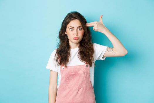 Image of tired and shocked young woman pointing finger gun at head, blowing her mind, staring bothered at camera, standing against blue background.