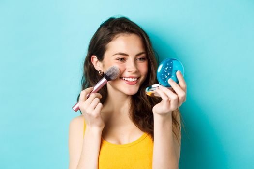 Beauty. Modern girl with candid smile applying make up with brush, looking in pocket mirror, standing over blue background.