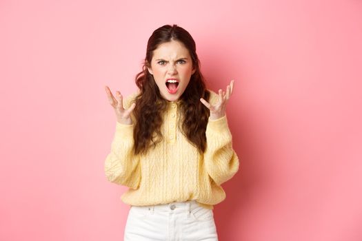 Angry and pissed-off girl screaming, shaking hands and grimacing furious, stare with outraged annoyed face at camera, standing against pink background.