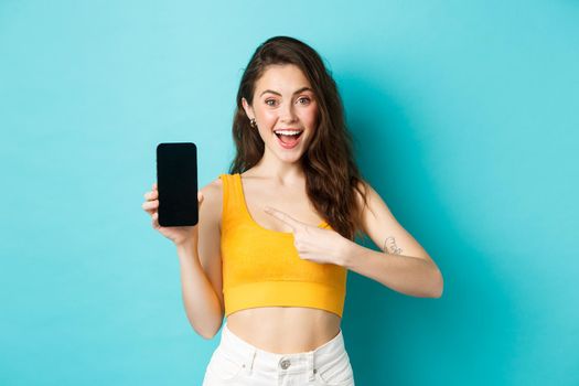 Check this out. Smiling young woman pointing at empty smartphone screen, recommending promo online, standing against blue background.