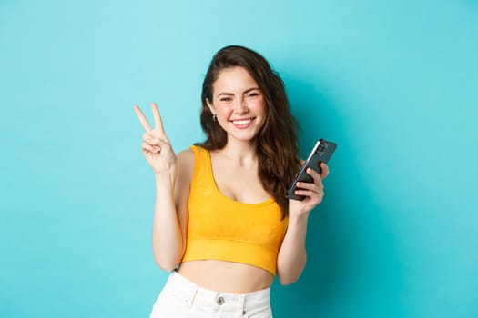 Technology and lifestyle concept. Beautiful cheerful girl sending positive vibes, smiling and showing peace sign, using cellphone, standing against blue background.