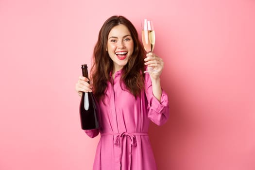 Celebration and holidays concept. Beautiful woman saying toast, raising glass of champagne and holding bottle, having fun at party, standing against pink background.
