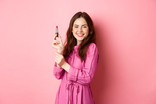 Coquettish smiling woman showing lipstick, applying make up for party event, standing in dress against pink background.