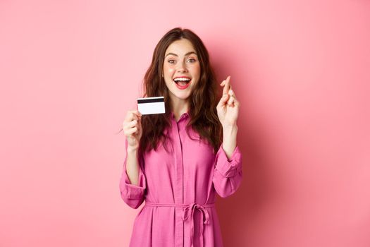 Image of hopeful young woman showing plastic credit card and cross fingers for good luck, making wish and smiling, standing against pink background. Copy space