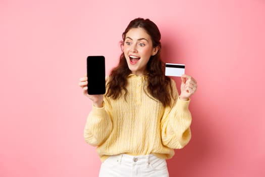 Technology and online shopping. Excited attractive girl showing smartphone screen, plastic credit card, look amazed at phone, standing against pink background.