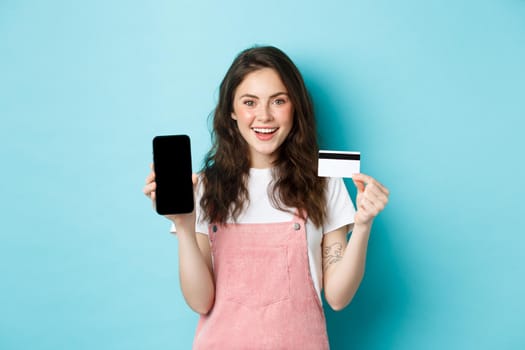 Image of beautiful smiling woman in stylish outfit, showing plastic credit card and empty smartphone screen, recommend app, standing over blue background.