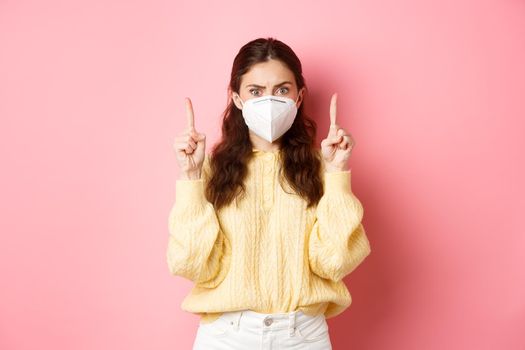 Covid, corona and social distancing concept. Confused and shocked young woman in medical respirator pointing fingers up, staring startled at camera, pink background.