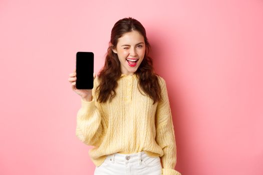 Image of cheeky attractive girl, showing you smartphone screen, winking and smiling, recommending mobile phone, standing against pink background.